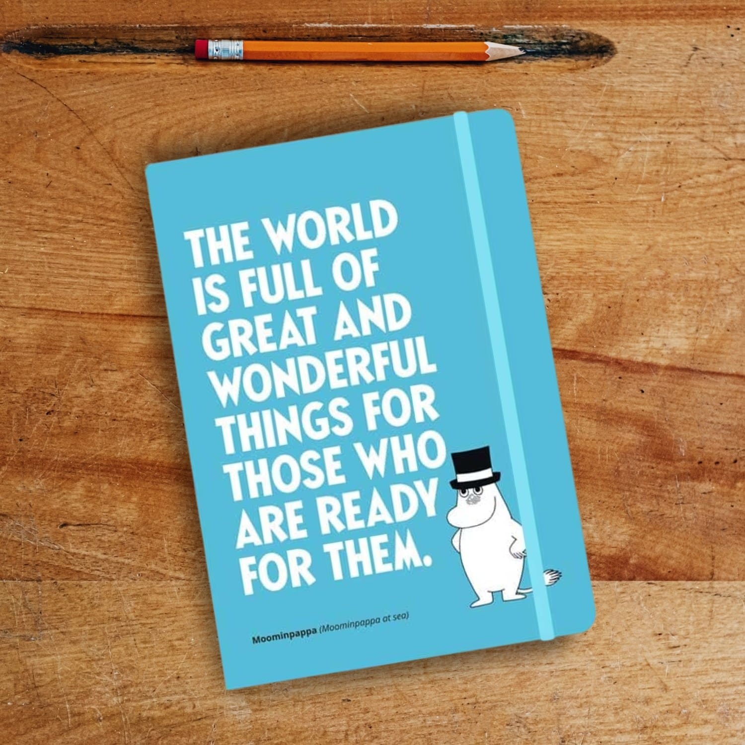 Ein blaues Mumin Notizbuch auf einem Holztisch. Darauf ist Muminpapa und ein englischer Spruch.